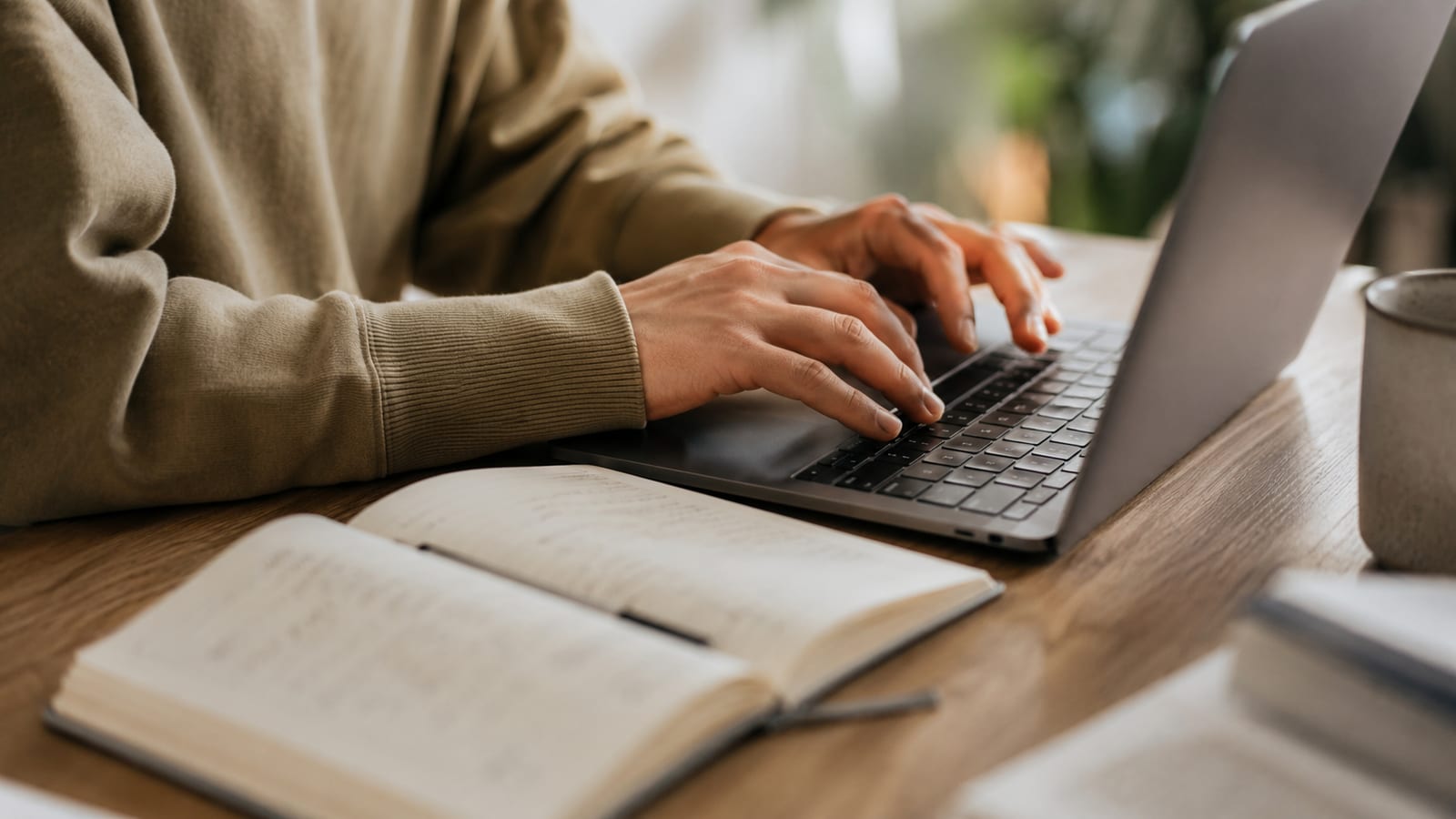 Primo piano di mani che digitano con cura su una tastiera, accanto un quaderno con appunti